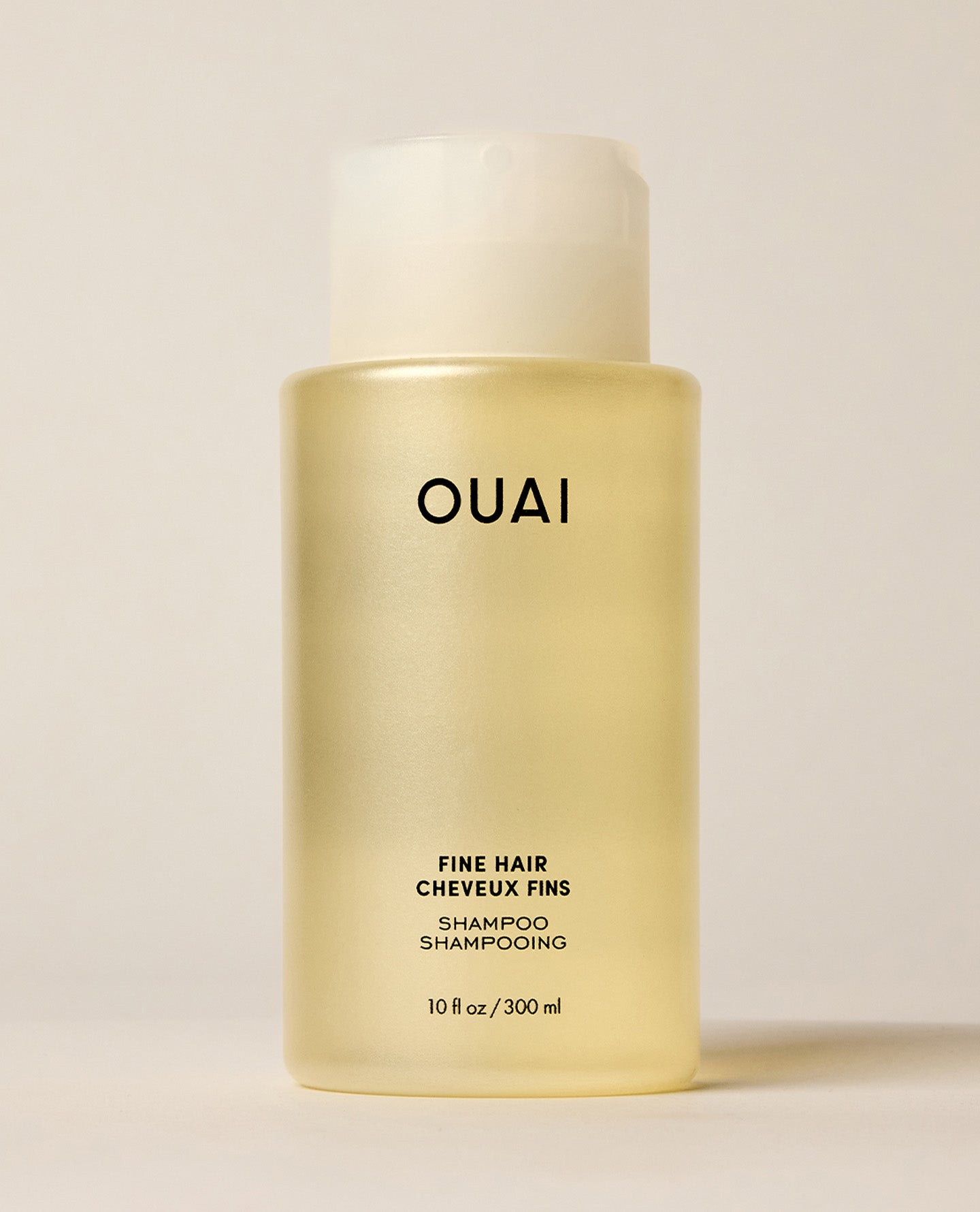 A translucent OUAI Fine Hair Shampoo bottle containing pale yellow liquid stands upright on a warm cream surface. Text: “OUAI”, “FINE HAIR”, “CHEVEUX FINS”, “SHAMPOO”, “SHAMPOOING”, “10 fl oz / 300 ml”.