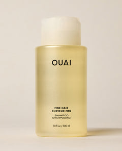 A translucent OUAI Fine Hair Shampoo bottle containing pale yellow liquid stands upright on a warm cream surface. Text: “OUAI”, “FINE HAIR”, “CHEVEUX FINS”, “SHAMPOO”, “SHAMPOOING”, “10 fl oz / 300 ml”.