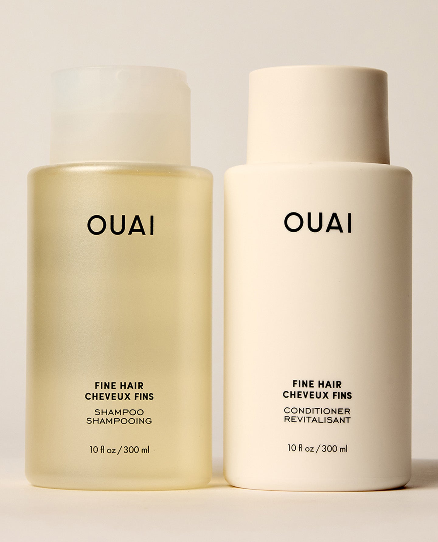 Two OUAI Fine Hair shampoo and conditioner bottles stand side by side on a cream studio surface, comprising a translucent yellow shampoo bottle and opaque ivory conditioner bottle.