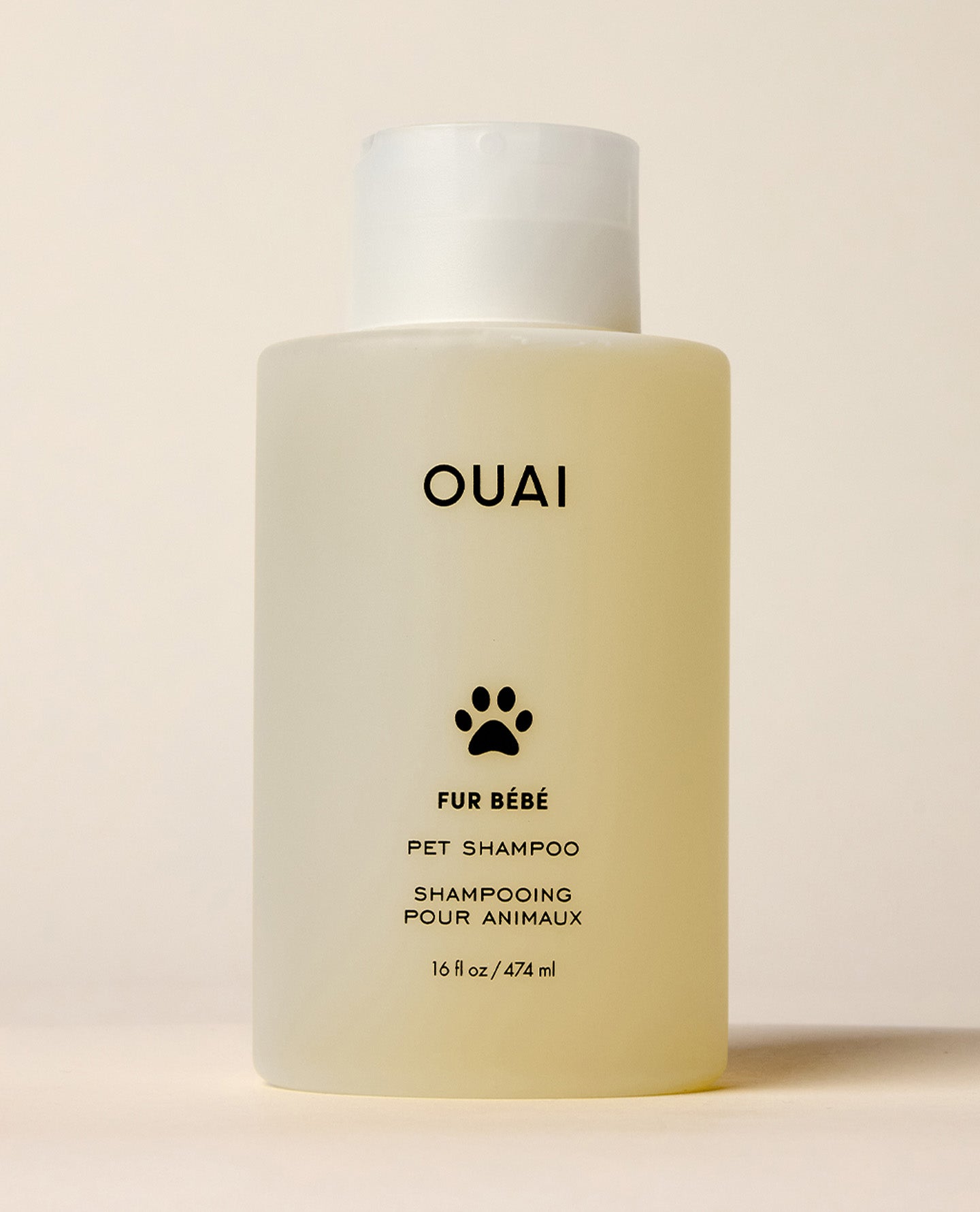 A translucent OUAI Fur Bébé Pet Shampoo bottle containing pale yellow liquid stands against a cream studio background. Text: “OUAI”, “FUR BÉBÉ”, “PET SHAMPOO”, “SHAMPOOING POUR ANIMAUX”, “16 fl oz / 474 ml”.
