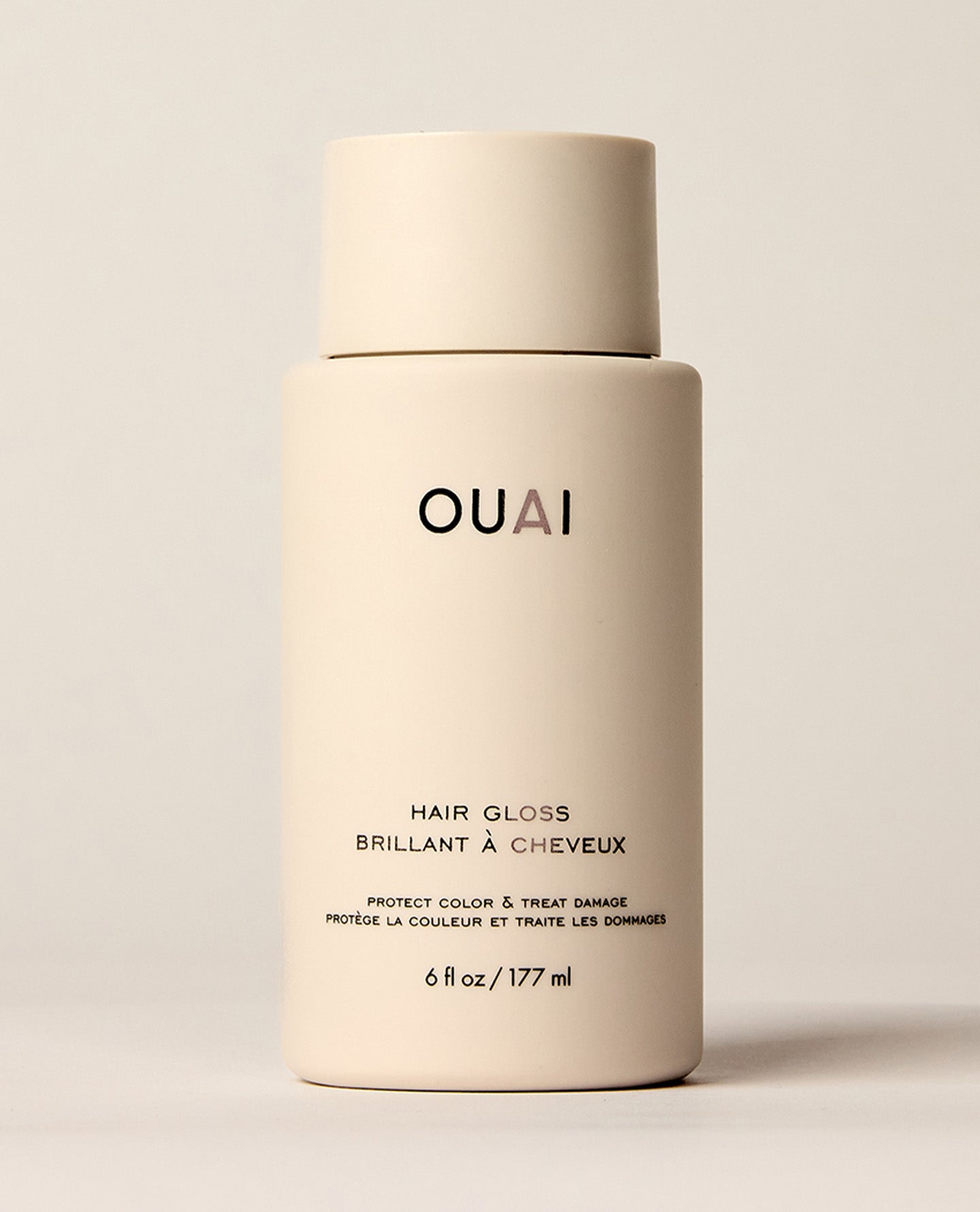 An ivory OUAI Hair Gloss bottle stands upright against a warm neutral background. Text: “OUAI”, “HAIR GLOSS”, “BRILLANT À CHEVEUX”, “PROTECT COLOR & TREAT DAMAGE”, “PROTÈGE LA COULEUR ET TRAITE LES DOMMAGES”, “6 fl oz / 177 ml”.