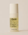 A small cylindrical OUAI Hair Oil bottle containing pale golden liquid stands upright on a cream surface, topped with a matte white cap.
