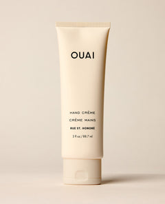 An ivory OUAI hand cream tube stands upright on a warm cream studio surface. Text: “OUAI”, “HAND CRÈME”, “CRÈME MAINS”, “RUE ST. HONORÉ”, “3 fl oz / 88.7 ml”.
