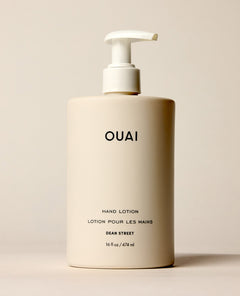 An ivory OUAI Dean Street Hand Lotion pump bottle stands upright on a softly lit cream surface. Text: “OUAI”, “HAND LOTION”, “LOTION POUR LES MAINS”, “DEAN STREET”, “16 fl oz / 474 ml”.