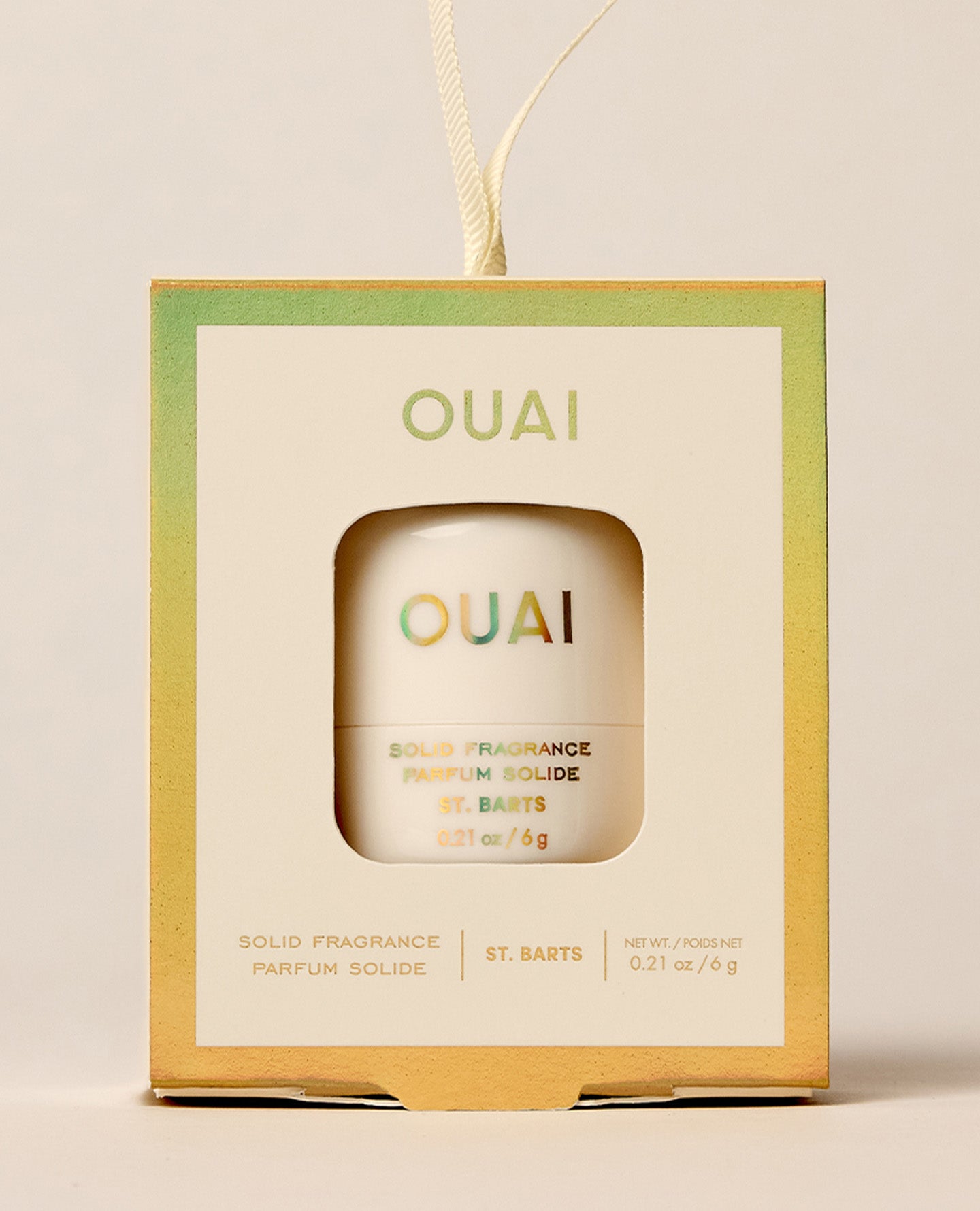 A boxed OUAI St. Barts solid fragrance ornament hangs by a cream cord, with the compact visible through a square window against a warm neutral backdrop.