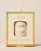 A boxed OUAI St. Barts solid fragrance ornament hangs by a cream cord, with the compact visible through a square window against a warm neutral backdrop.