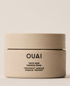 A wide beige OUAI Thick Hair Treatment Masque jar sits on a cream studio surface, tightly framed against a softly lit neutral background.