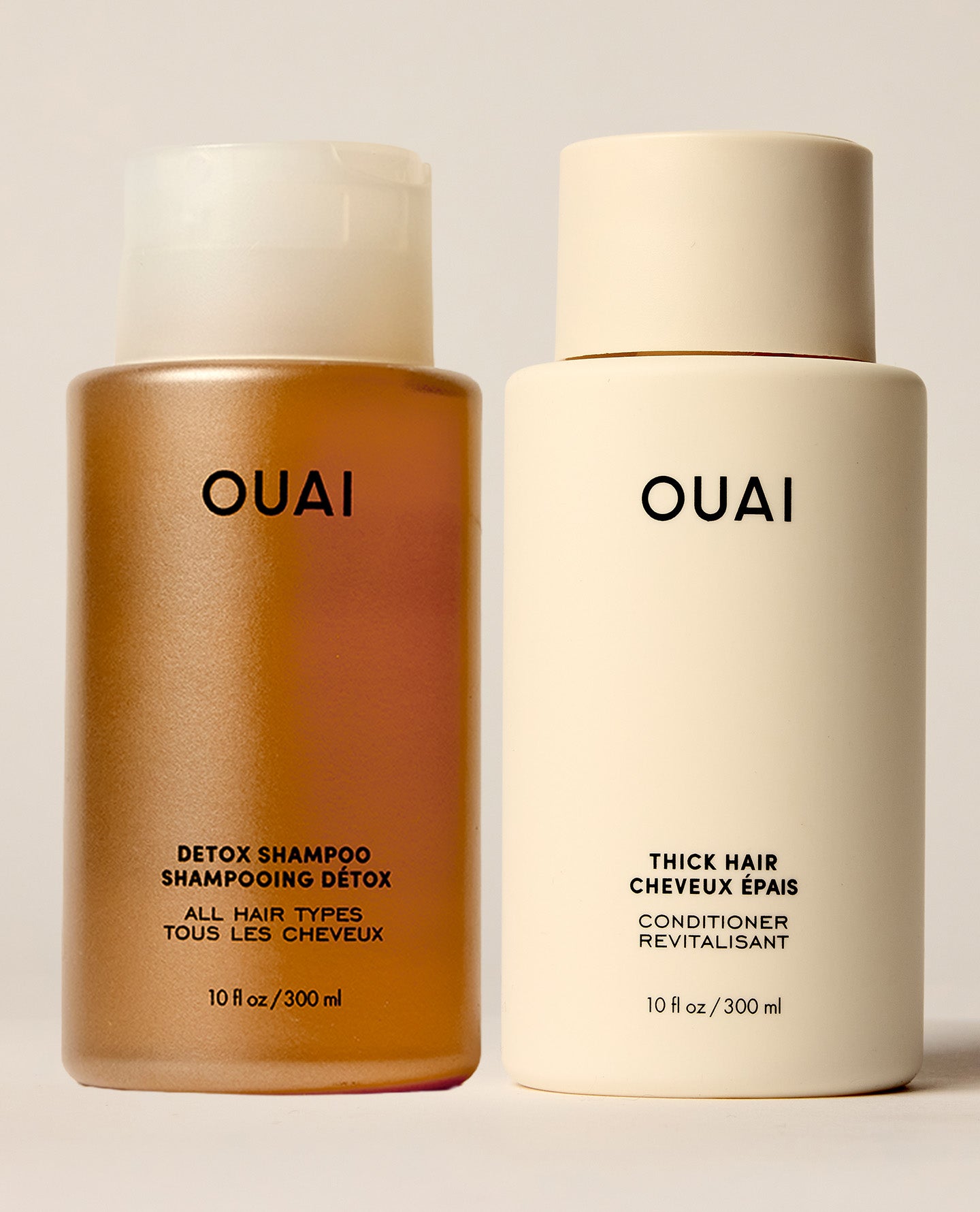 Two OUAI hair care bottles on a light beige background: a metallic amber bottle of detox shampoo and a matte cream bottle of thick hair conditioner.