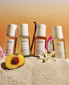 Four OUAI fragrance mist bottles arranged on sand with decorative flowers and fruit slices, against a warm beige-to-orange gradient background.