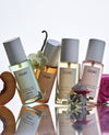 Four OUAI fragrance mist bottles arranged on a reflective surface with fruit and flower props against a clean white background. The bottles contain pale green, peach, yellow, and pink liquids and are labeled with OUAI.