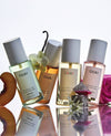 Four OUAI fragrance mist bottles arranged on a glossy reflective surface with fruit and flower props around them. The bottles contain pale green, peach, yellow, and blush-toned liquids with white caps and white labels showing the OUAI brand.