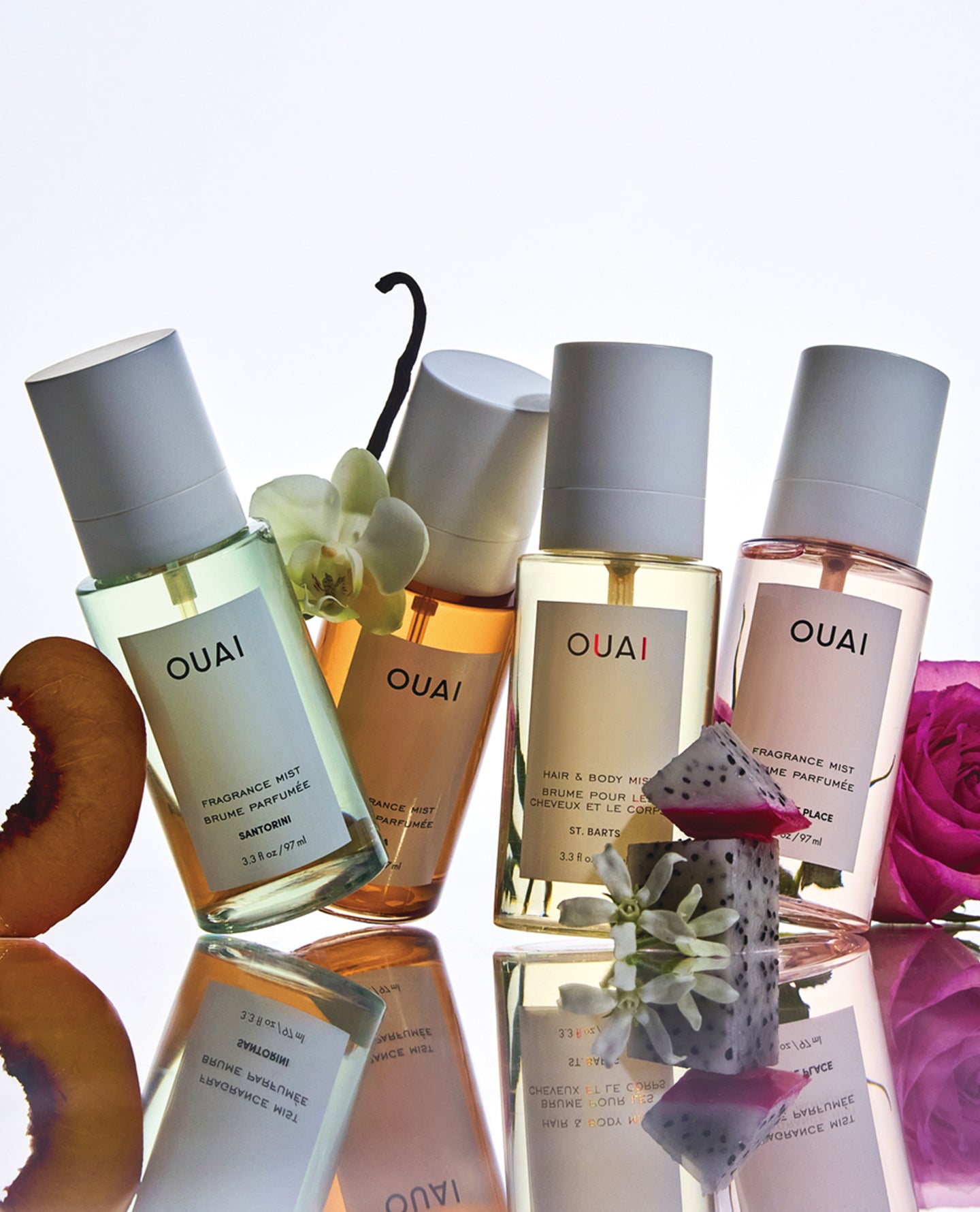 Four OUAI fragrance mist bottles arranged on a glossy reflective surface with fruit and flower props around them. The bottles contain pale green, peach, yellow, and blush-toned liquids with white caps and white labels showing the OUAI brand.