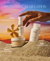 Three OUAI skincare products arranged in sand with a hand reaching toward the bottle, against a sunset sky background. Text labels identify Step 1 Body Cleanser, Step 2 Body Crème, and Step 3 Hair & Body Mist.