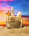 OUAI St. Bart's beach-themed skincare set arranged in a clear zippered pouch filled with sand, with a gold frame, set against a vibrant beach sunset sky and sand dunes.