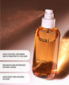 A bottle of OUAI Wave Spray with a white pump dispenser, shown spraying a fine mist. The bottle is filled with a golden-amber liquid and features black text.