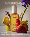 A bottle of OUAI North Bondi Fragrance wave spray stands centered against a warm beige background, surrounded by fruit and flowers including a halved lemon, raspberries, a red apple, a cinnamon stick, and purple and pink blossoms. Text at the top reads 'NORTH BONDI FRAGRANCE' and text at the bottom reads 'NOTES OF: ITALIAN LEMON, BERGAMOT, JASMINE & VIOLET'.