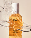 A bottle of OUAI Wave Spray oil with golden amber liquid, shown with a dynamic water splash and droplet effect against a light beige background.