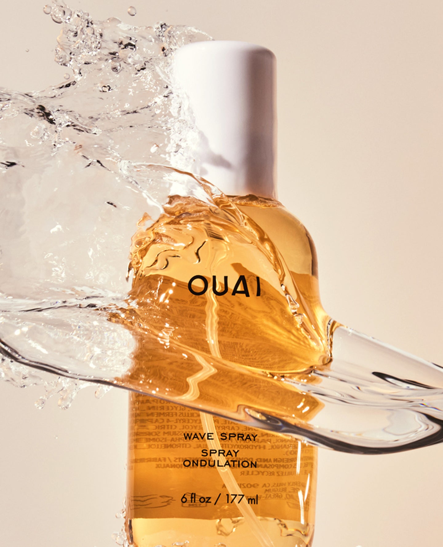 A bottle of OUAI Wave Spray oil with golden amber liquid, shown with a dynamic water splash and droplet effect against a light beige background.
