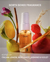 A bottle of OUAI North Bondi Fragrance spray centered among fruit and botanical props, including a halved lime, raspberries, cinnamon sticks, wheat, and a pink rose, with text reading 'NORTH BONDI FRAGRANCE' and 'NOTES OF: ITALIAN LEMON, BERGAMOT, JASMINE & VIOLET'.