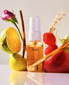 A bottle of OUAI wave spray with amber liquid and a white spray top, styled with citrus halves, raspberries, cinnamon sticks, wheat, and a rose on a clean light background.