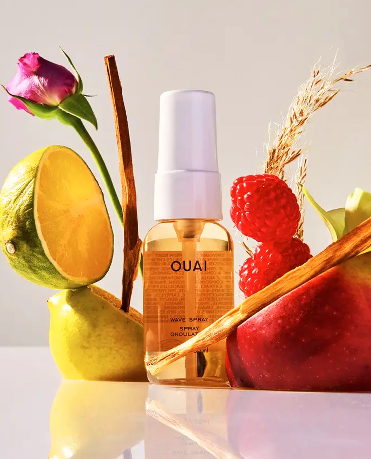 A bottle of OUAI wave spray with amber liquid and a white spray top, styled with citrus halves, raspberries, cinnamon sticks, wheat, and a rose on a clean light background.