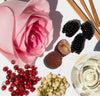 Pink rose, blackberries, lychees, red currants, dried herbs, and a glass of white wine on a white surface.