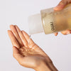 A clear bottle labeled OUAI pours a transparent gel cleanser onto an open hand against a clean white background.